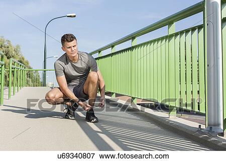 Stock Photo - Full length of sporty man tying shoelace on bridge. Fotosearch