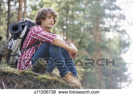Stock Photo - Full length of thoughtful male backpacker relaxing on cliff in forest. Fotosearch