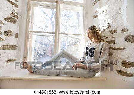 Full length of thoughtful young woman sitting on window sill at home View Large Photo Image Stock Photo - Full length of thoughtful young woman sitting on window sill at home. Fotosearch