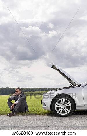 Stock Image - Full length of unhappy young businessman sitting by broken down car at countryside. Fotosearch