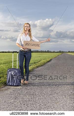 Stock Photo - Full length of woman hitching while holding anywhere sign on country road. Fotosearch