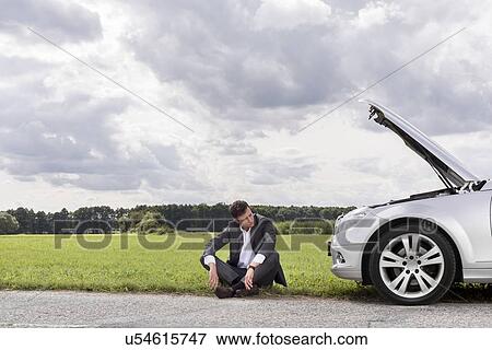 Stock Photo - Full length of worried businessman sitting by broken down car at countryside. Fotosearch