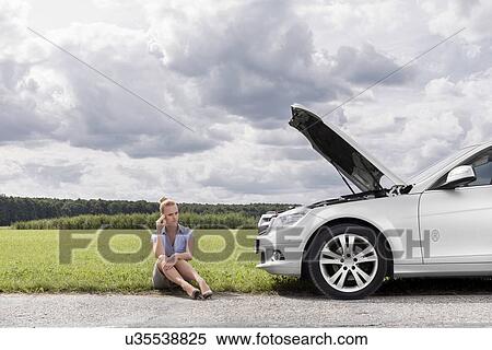 Stock Photography - Full length of worried businesswoman sitting by broken down car at countryside. Fotosearch