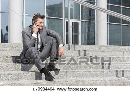 Picture - Full length of young businessman answering cell phone on steps outside office. Fotosearch