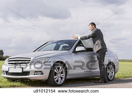 Picture - Full length of young businessman reading map by car at countryside. Fotosearch