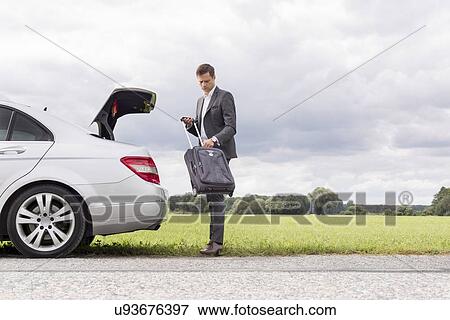 Stock Photo - Full length of young businessman unloading suitcase from broken down car at countryside. Fotosearch