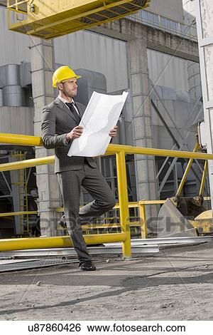 Stock Photograph - Full length of young businessman examining blueprint at construction site. Fotosearch