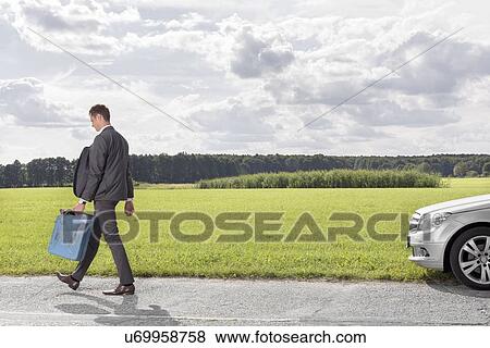 Stock Photo - Full length of young businessman with gas can leaving broken down car at countryside. Fotosearch