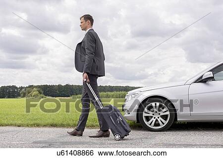 Stock Photograph - Full length of young businessman with suitcase walking by broken down car at countryside. Fotosearch
