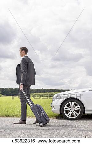 Full length of young businessman with suitcase leaving broken down car at countryside View Large Photo Image Stock Photograph - Full length of young businessman with suitcase leaving broken down car at countryside. Fotosearch
