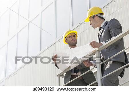 Stock Photo - Full length of young businessmen discussing over blueprint on stairway. Fotosearch