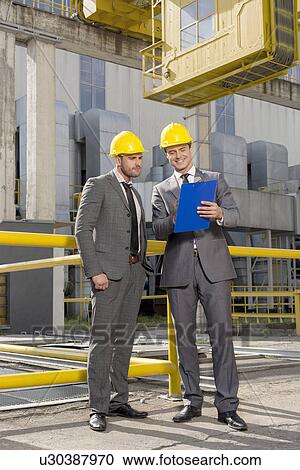 Stock Image - Full length of young businessmen with clipboard discussing at construction site. Fotosearch