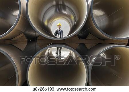 Stock Image - Full length of young male architect through stacked pipes at construction site. Fotosearch