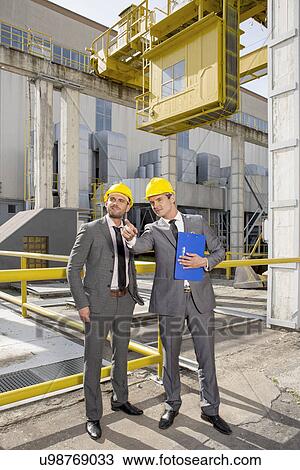 Stock Image - Full length of young male architect with colleague pointing away at construction site. Fotosearch