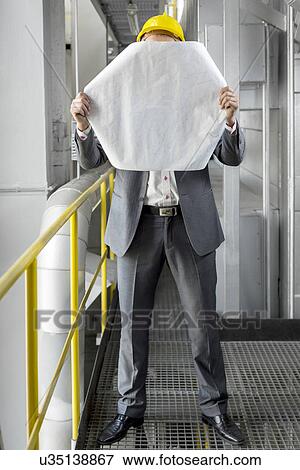 Full length of young male architect holding blueprint in front of face at industry View Large Photo Image Stock Photo - Full length of young male architect holding blueprint in front of face at industry. Fotosearch