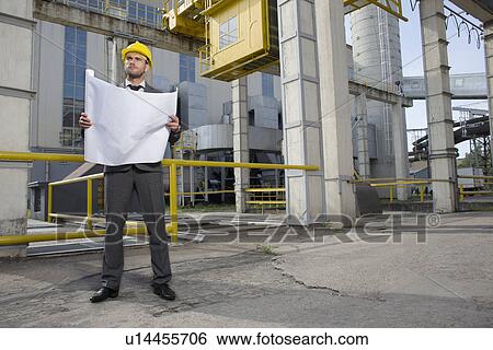 Stock Photograph - Full length of young male architect with blueprint outside industry. Fotosearch
