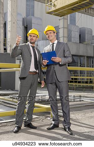 Stock Image - Full length of young male architects with clipboard discussing outside industry. Fotosearch