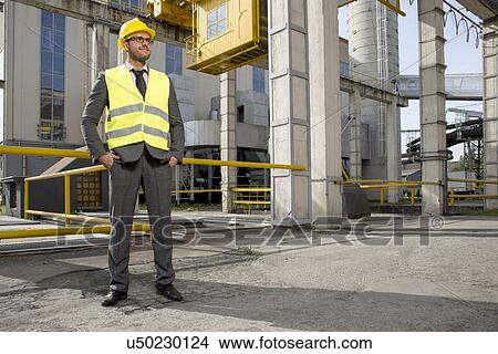 Full length of young male engineer with hands in pockets standing outside industry View Large Photo Image Picture - Full length of young male engineer with hands in pockets standing outside industry. Fotosearch