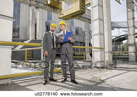 Stock Image - Full length of young male engineer with clipboard looking away at construction site. Fotosearch