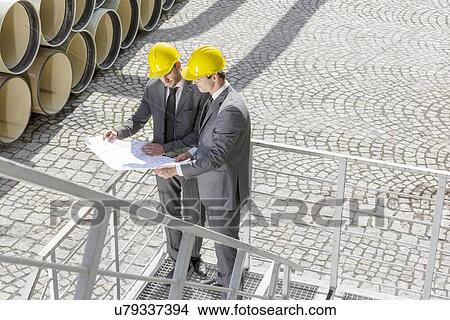 Picture - Full length of young male engineers examining blueprint on stairway. Fotosearch