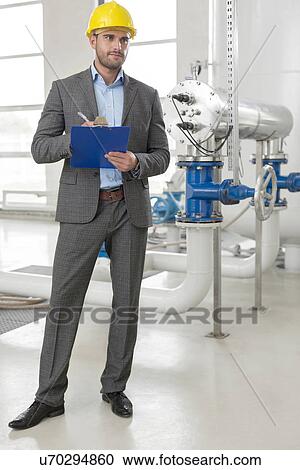 Stock Image - Full length of young male supervisor writing on clipboard by industrial machinery. Fotosearch