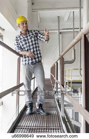 Stock Photo - Full length of young male worker gesturing on metal aisle in industry. Fotosearch