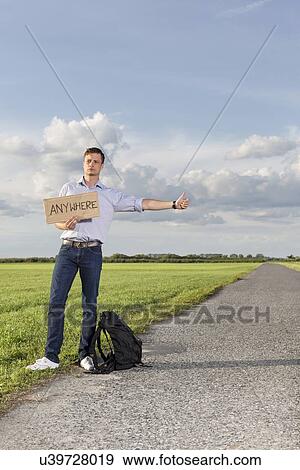 Stock Photo - Full length of young man hitching while holding anywhere sign on countryside. Fotosearch