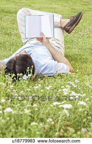 Stock Image - Full length of young man holding book while lying on grass in park. Fotosearch