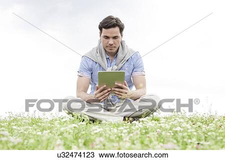 Full length of young man holding digital tablet while sitting on grass against sky View Large Photo Image Stock Image - Full length of young man holding digital tablet while sitting on grass against sky. Fotosearch