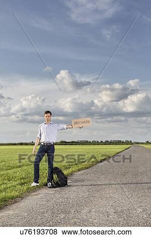 Stock Photo - Full length of young man holding anywhere sign on empty road. Fotosearch