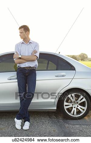 Picture - Full length of young man looking away while standing by car at countryside. Fotosearch