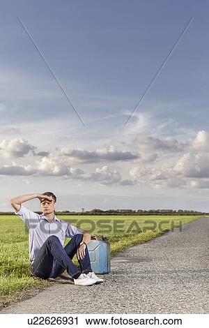 Full length of young man shielding eyes while sitting with petrol can on country road View Large Photo Image Stock Image - Full length of young man shielding eyes while sitting with petrol can on country road. Fotosearch