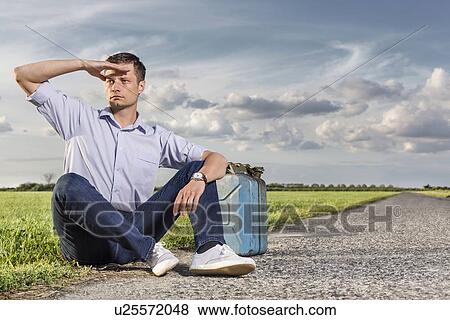 Full length of young man shielding eyes while sitting with petrol can by country road View Large Photo Image Stock Photo - Full length of young man shielding eyes while sitting with petrol can by country road. Fotosearch