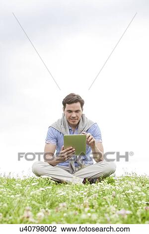 Full length of young man using digital tablet while sitting on grass against sky View Large Photo Image Stock Image - Full length of young man using digital tablet while sitting on grass against sky. Fotosearch