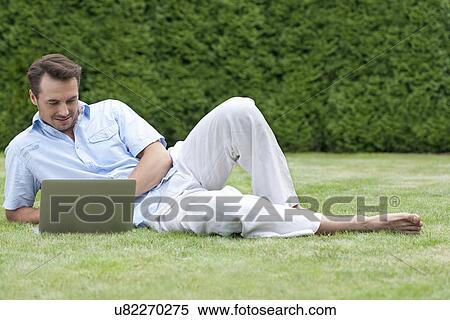 Stock Photography - Full length of young man using laptop while lying on grass in park. Fotosearch