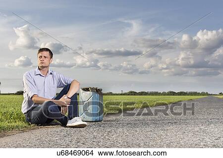 Picture - Full length of young man with empty gas can sitting by country road. Fotosearch