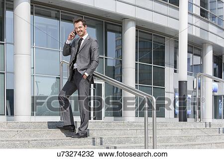 Stock Image - Full length portrait of smiling businessman answering cell phone while standing on steps outside office. Fotosearch