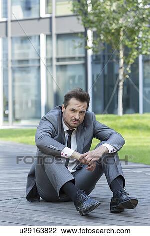 Stock Image - Full length portrait of stressed businessman sitting on path outside office. Fotosearch