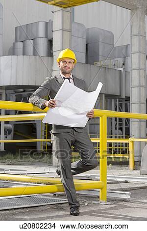 Picture - Full length portrait of young male architect with blueprint outside industry. Fotosearch