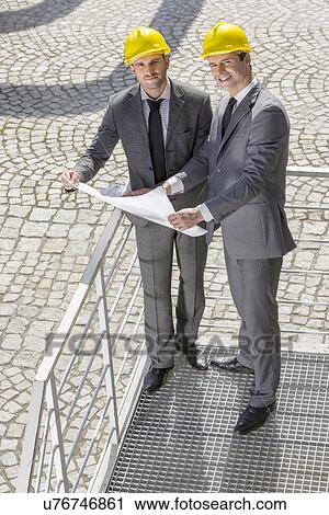 Stock Image - Full length portrait of young businessmen with blueprint on stairway. Fotosearch