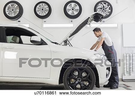 Stock Image - Full length side view of male mechanic examining car engine in repair shop. Fotosearch
