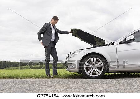 Picture - Full length side view of young businessman examining broken down car engine at countryside. Fotosearch