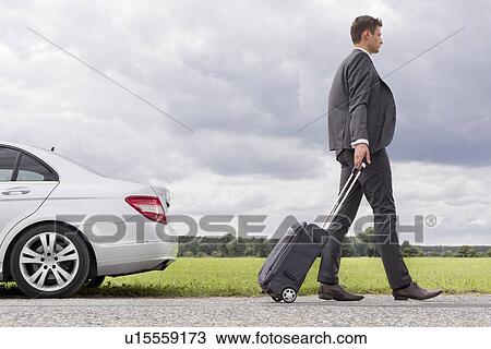 Stock Image - Full length side view of young businessman with luggage leaving broken down car at countryside. Fotosearch
