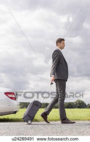 Stock Image - Full length side view of young businessman with luggage leaving broken down car at countryside. Fotosearch