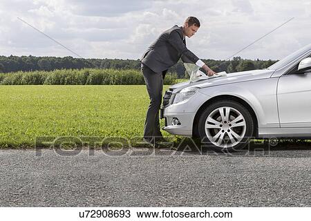 Stock Image - Full length side view of young businessman reading map on car hood at countryside. Fotosearch