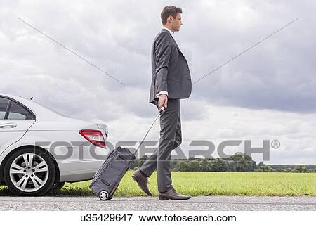 Stock Photo - Full length side view of young businessman with luggage leaving broken down car at countryside. Fotosearch