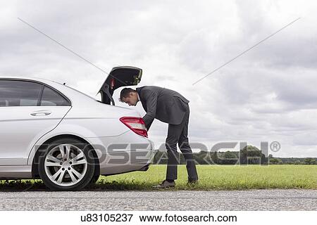 Stock Photo - Full length side view of young businessman looking in car trunk at countryside. Fotosearch