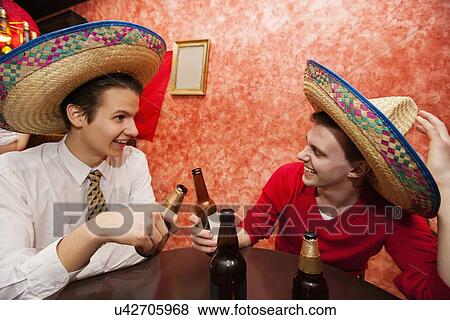 Stock Photo - Happy friends wearing Mexican hats toasting at restaurant table. Fotosearch