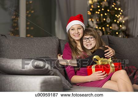 Stock Photo - Happy girl hugging sister on sofa during Christmas. Fotosearch