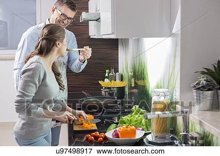 Happy man feeding food to woman cutting vegetables in kitchen View Large Photo Image Stock Photo - Happy man feeding food to woman cutting vegetables in kitchen. Fotosearch